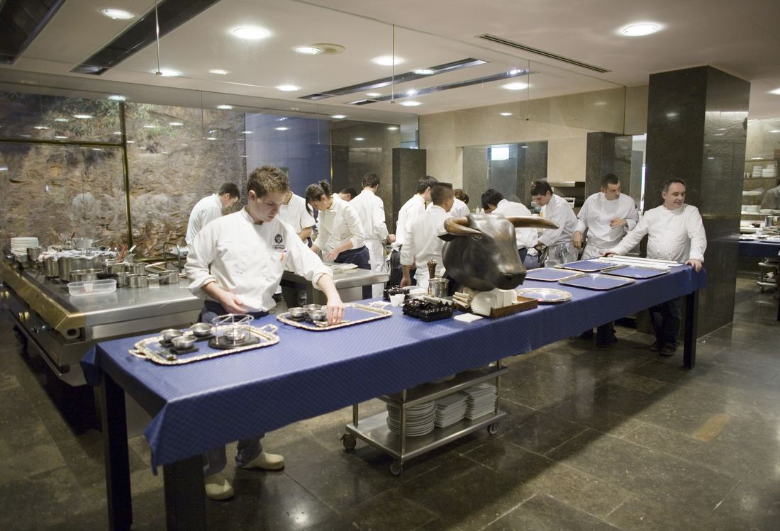 Chefs in white working at a long table in the bright kitchen of El Bulli